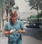 Brennon on a sidewalk somewhere in Germany looking as some sort of messy something to eat in his hands, chuckling