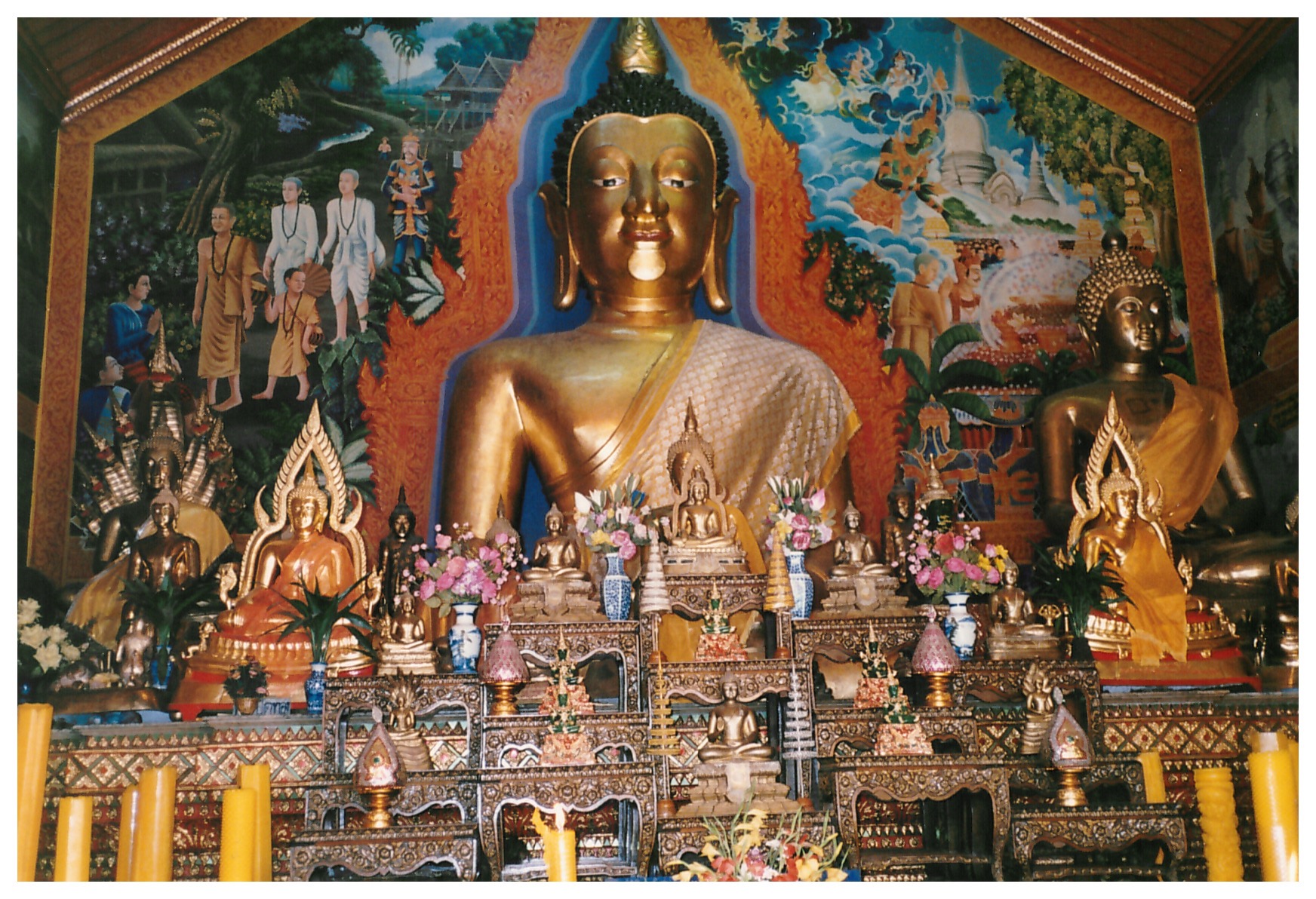 Dozens of small Buddha statues on ornate platforms in front of a large Buddha statue surrounded by a colorful mural
