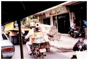 Two "mototrucks" in Bangkok traffic, one loaded with boxes and one with bundles of some sort of tubes