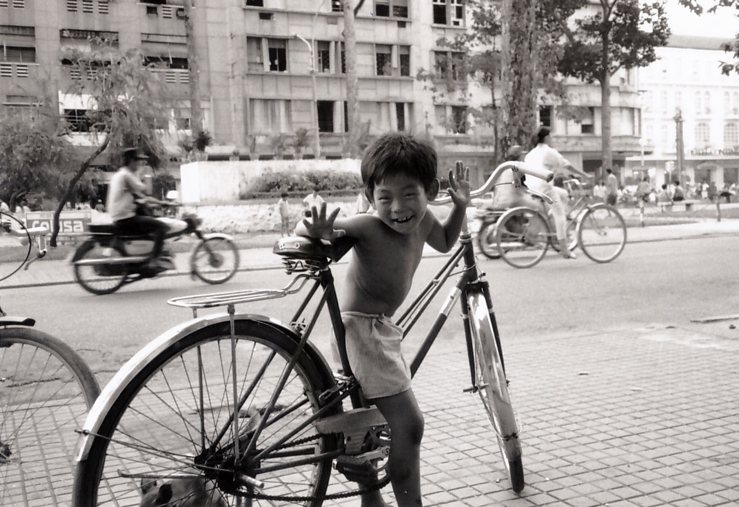 A young boy, straddling a bicycle far too large for him, mugs for the camera as bicycles and motorcycles speed by in the background