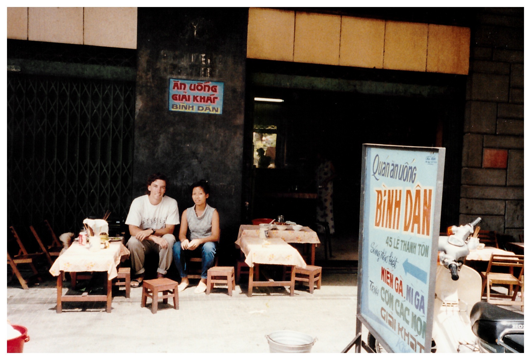 The author sitting on short stool at a street-side table with a young local woman