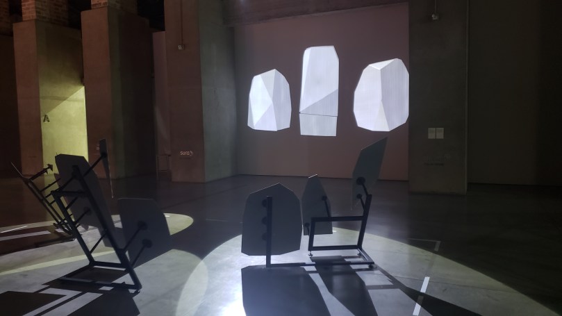 Artwork consisting of irregularly shaped mirrors mounted on metal floor stands, each of which are reflecting a spotlight onto the wall, resulting in precisely assembled reflections that combine to create three geometric shapes with various components of different shades of gray and white