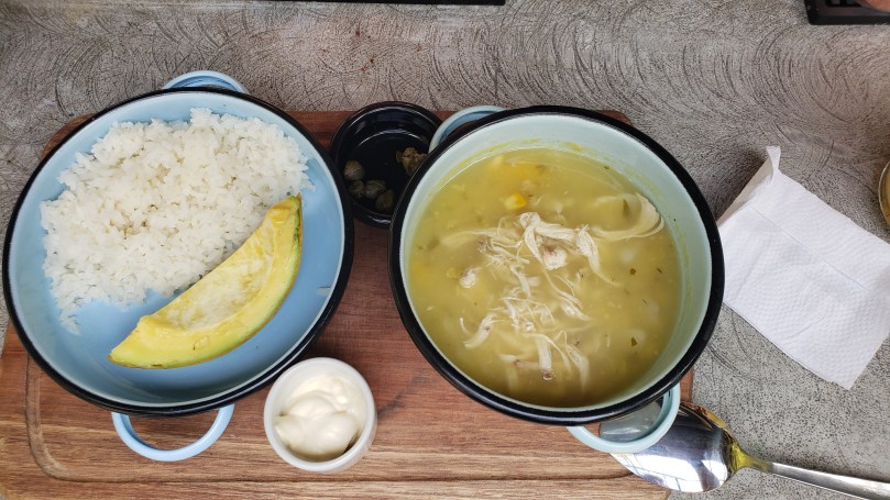 Two soup bowls - one with white rice and a slice of avocado and one with a chicken, potato, and corn stew soup. Also, two ramekins - one with a thick cream and one with a few capers. All served on a small wooden cutting board.