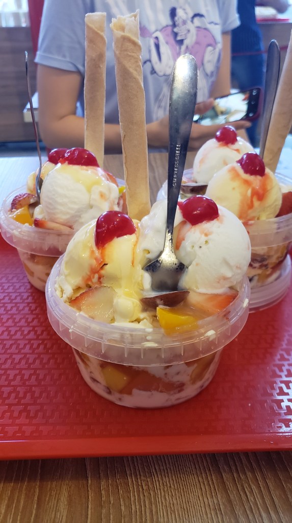 Three plastic bowls filled with fruit, sweetened condensed milk, and two scoops of vanilla ice cream, each with a spoon and a simple cylindrical cookie