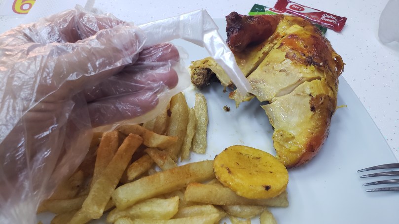 A hand in a clear plastic glove holding a piece of plastic that has been folded and tied into a knot over a plate of roasted chicken, french fries and a patacón