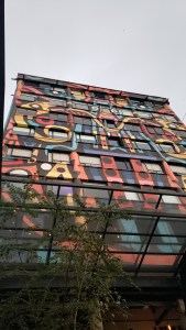 Looking up at an 8-story building completely covered in a brightly colored mural through open open glass-roofed atrium