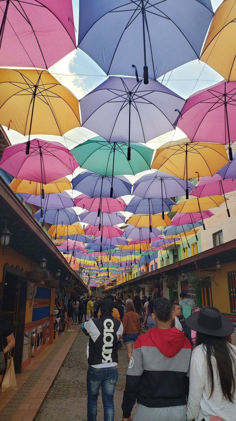 Beneath dozens (hundreds?) of colorful umbrellas, suspended 4-across, reaching from one building across the street to another along approximately 100 meters along a narrow pedestrian street