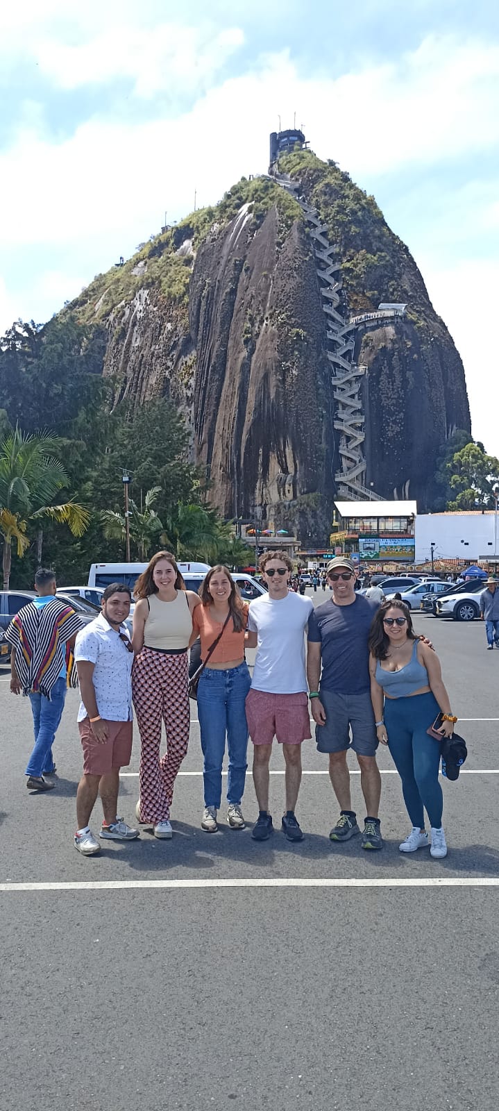 Three women and three men posing in a parking lot with a large, solitary rock in the background with a staircase zig zagging to the summit