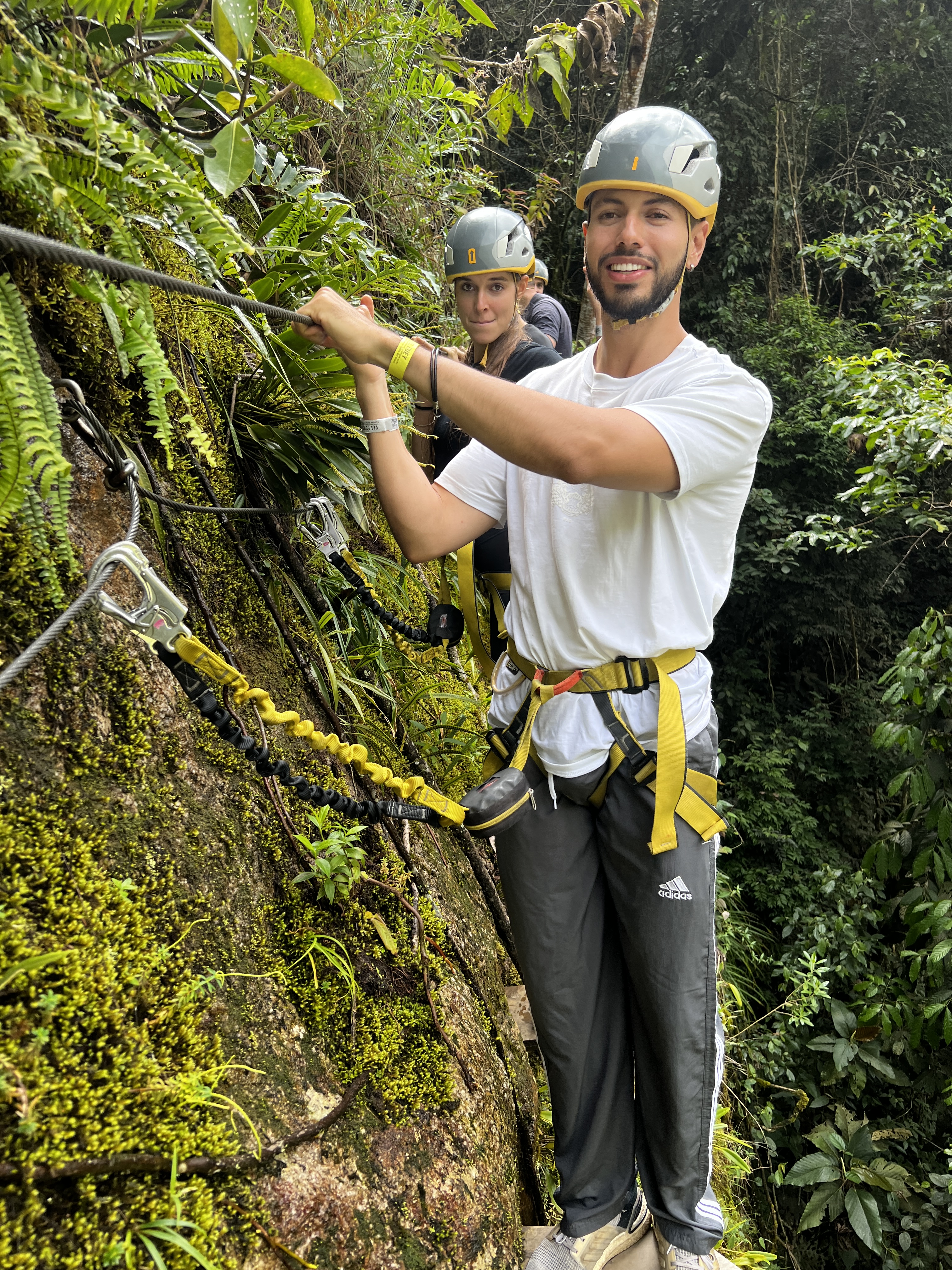 A man standing on the side of a cliff - helmeted, harnessed, and secured to the safety line - followed by other members of the group