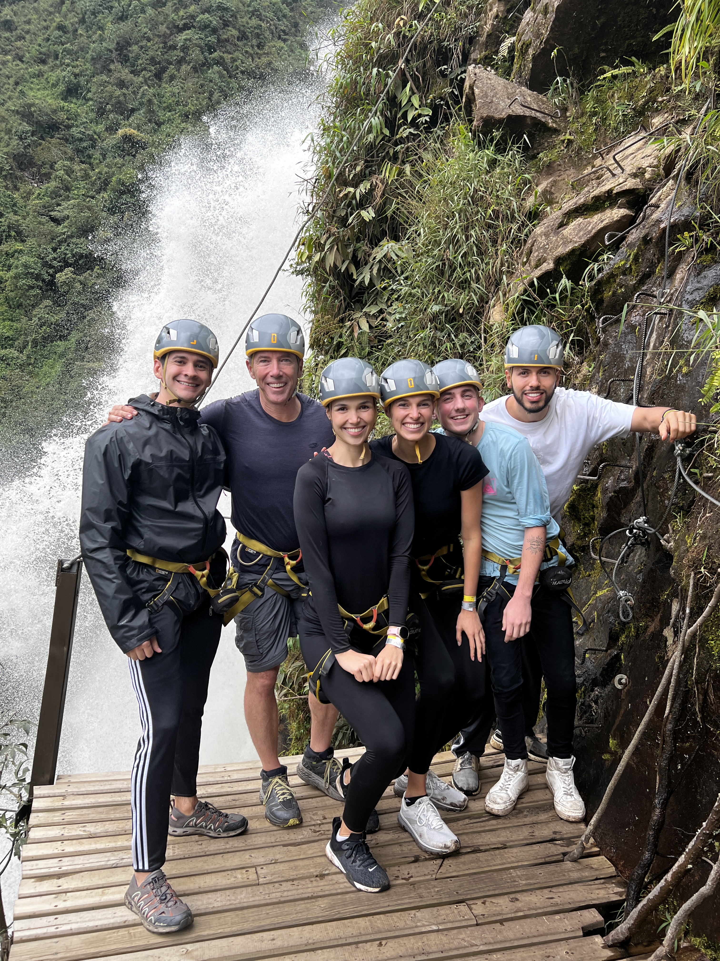 Two women and four men in helmets and harnesses, standing on a cliffside wooden platform