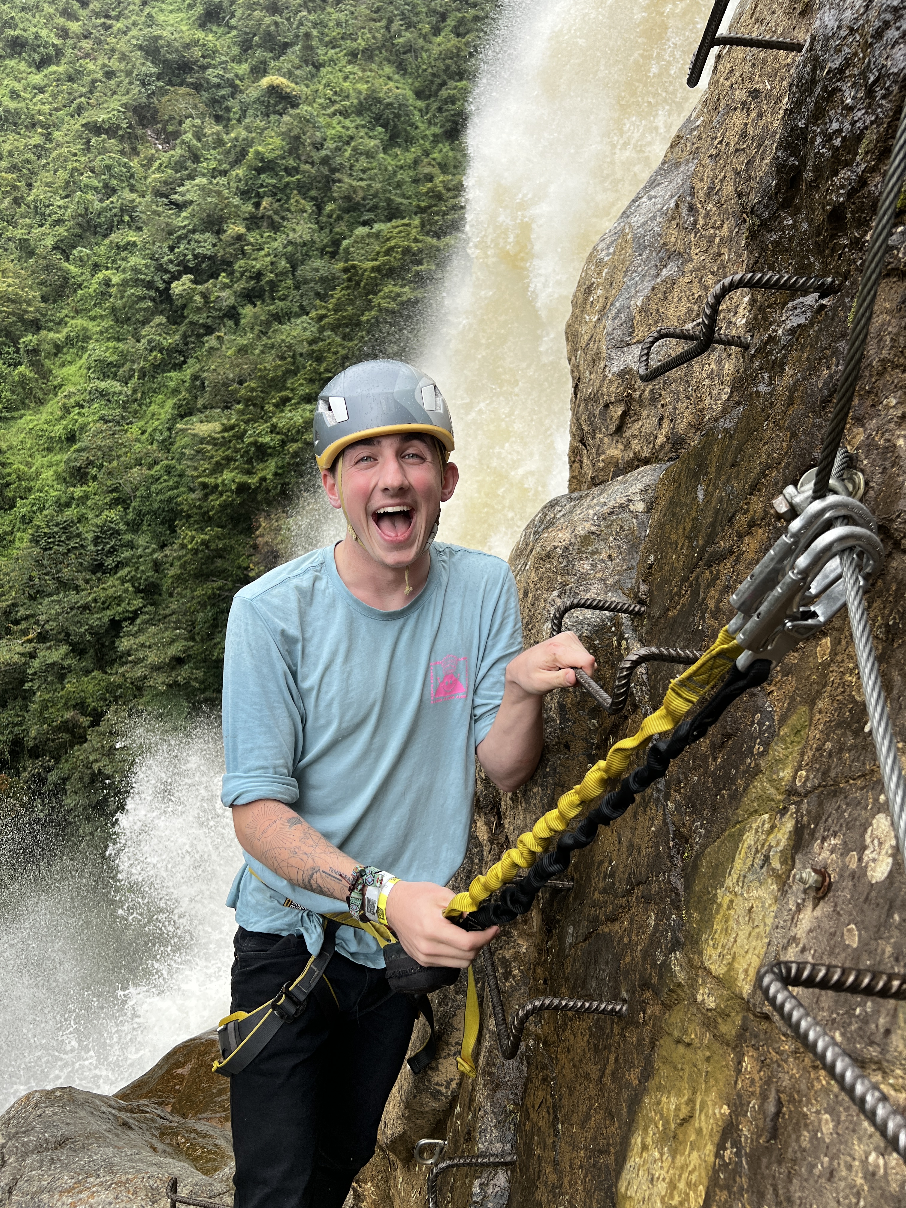 Helmeted and harnessed man, smiling with joy, attached to a metal safety line with the falls crashing down the cliff behind him