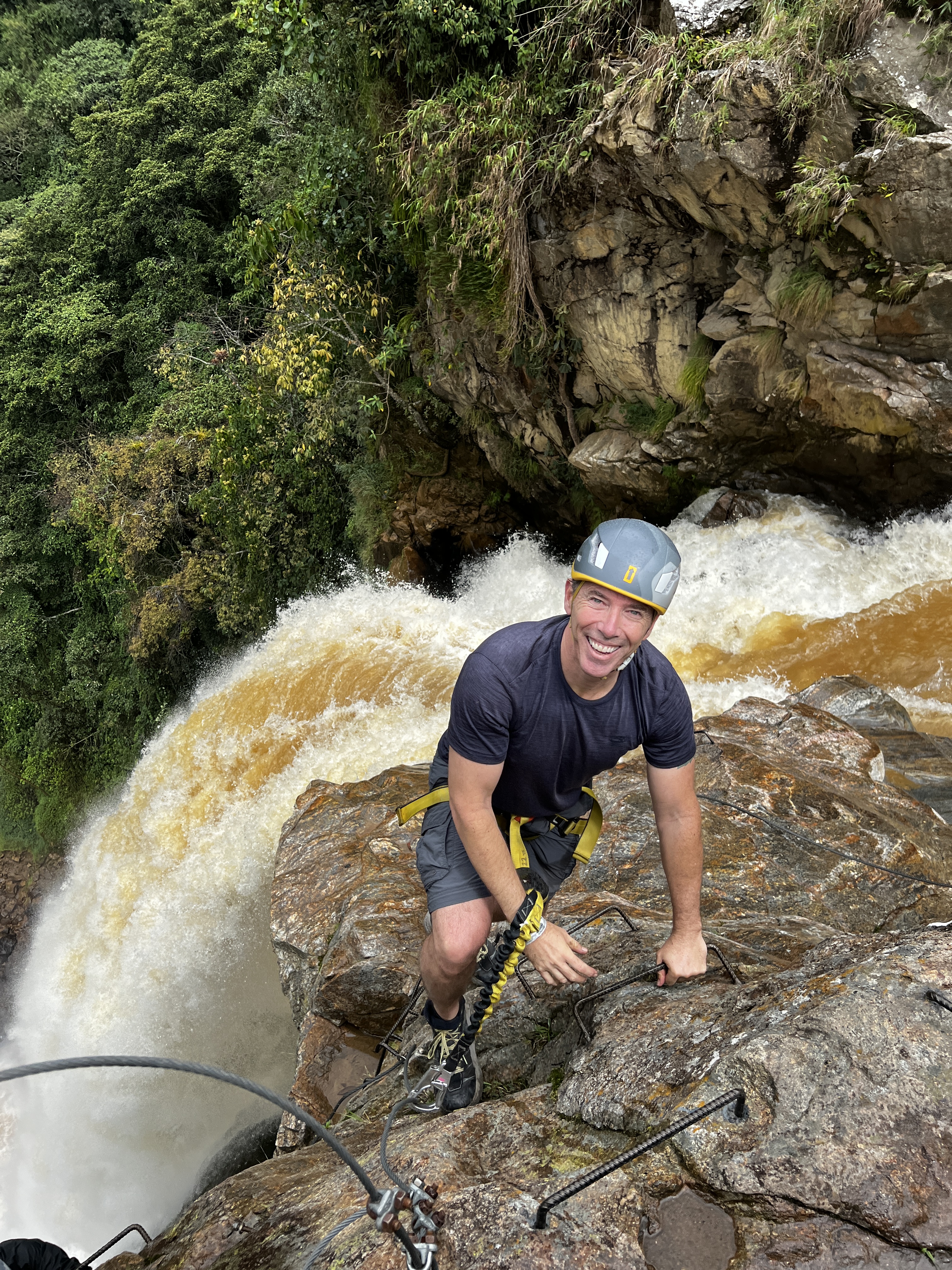 A smiling, helmeted and harnessed man on the crest of a cliff with the top of the waterfall just behind him