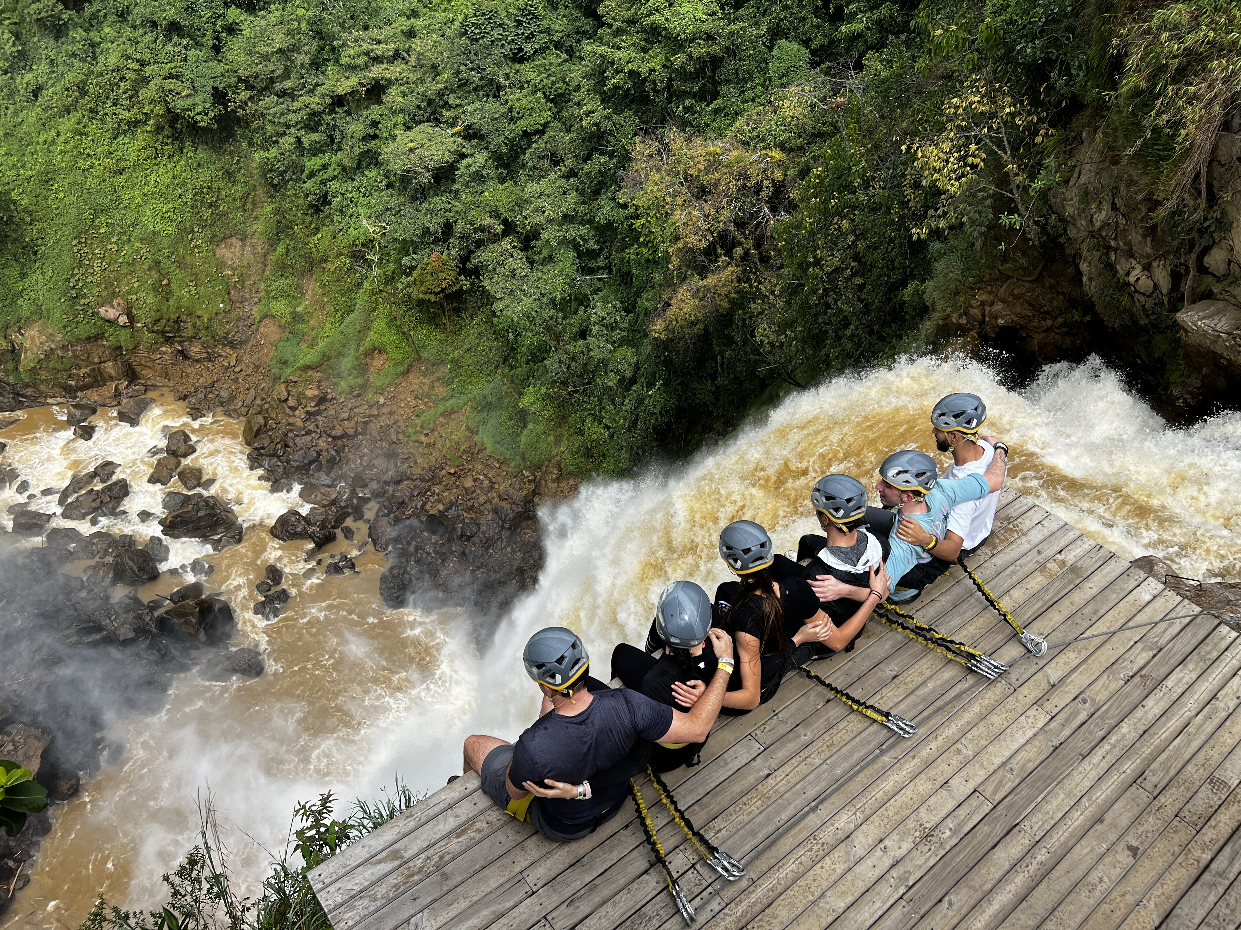 Six people arm in arm, sitting on a wood platform, dangling legs over the side, secured to a safety line