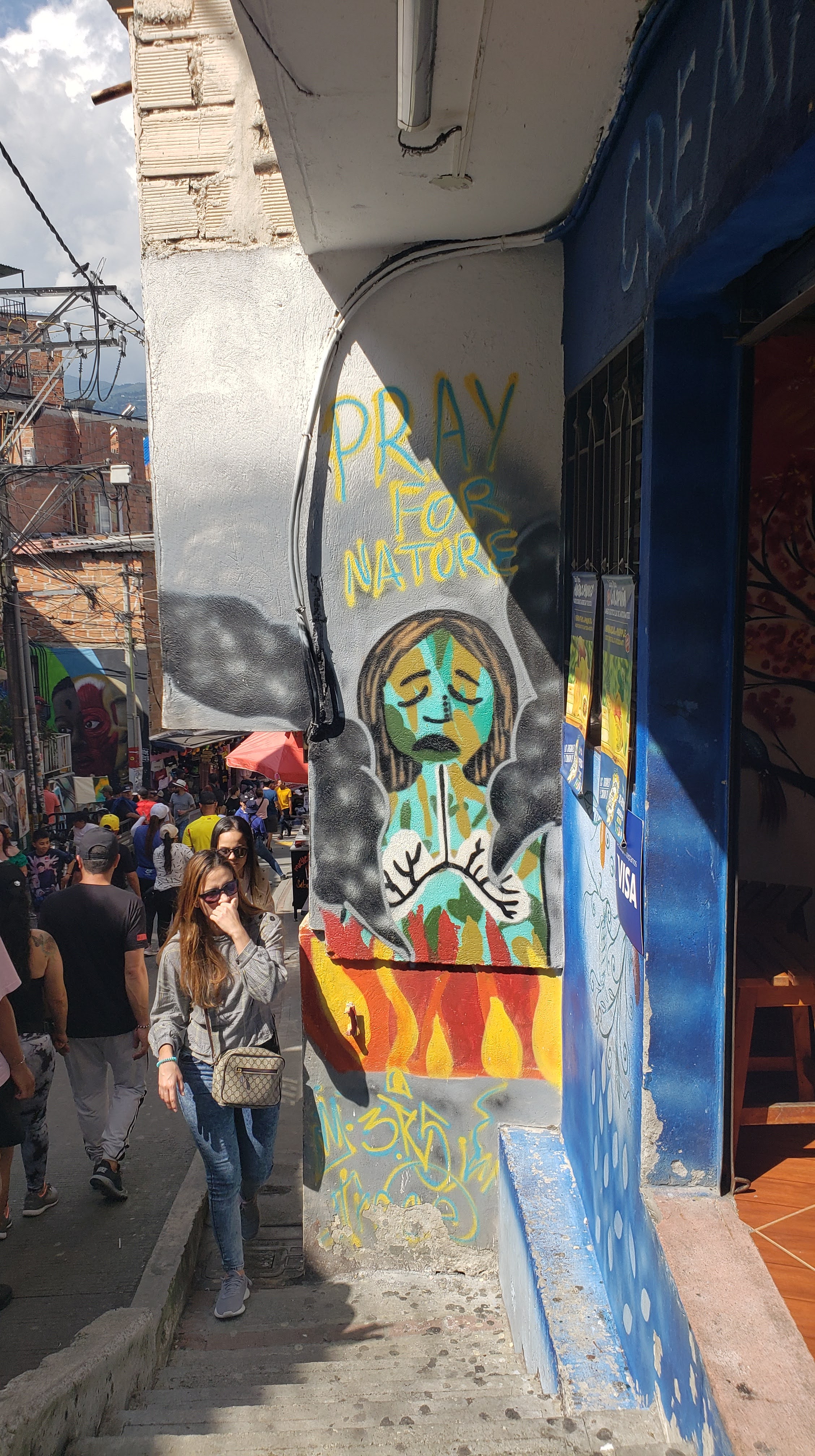 Small mural on a side wall at the base of some stairs that reads "Pray for Natore" above a weeping woman