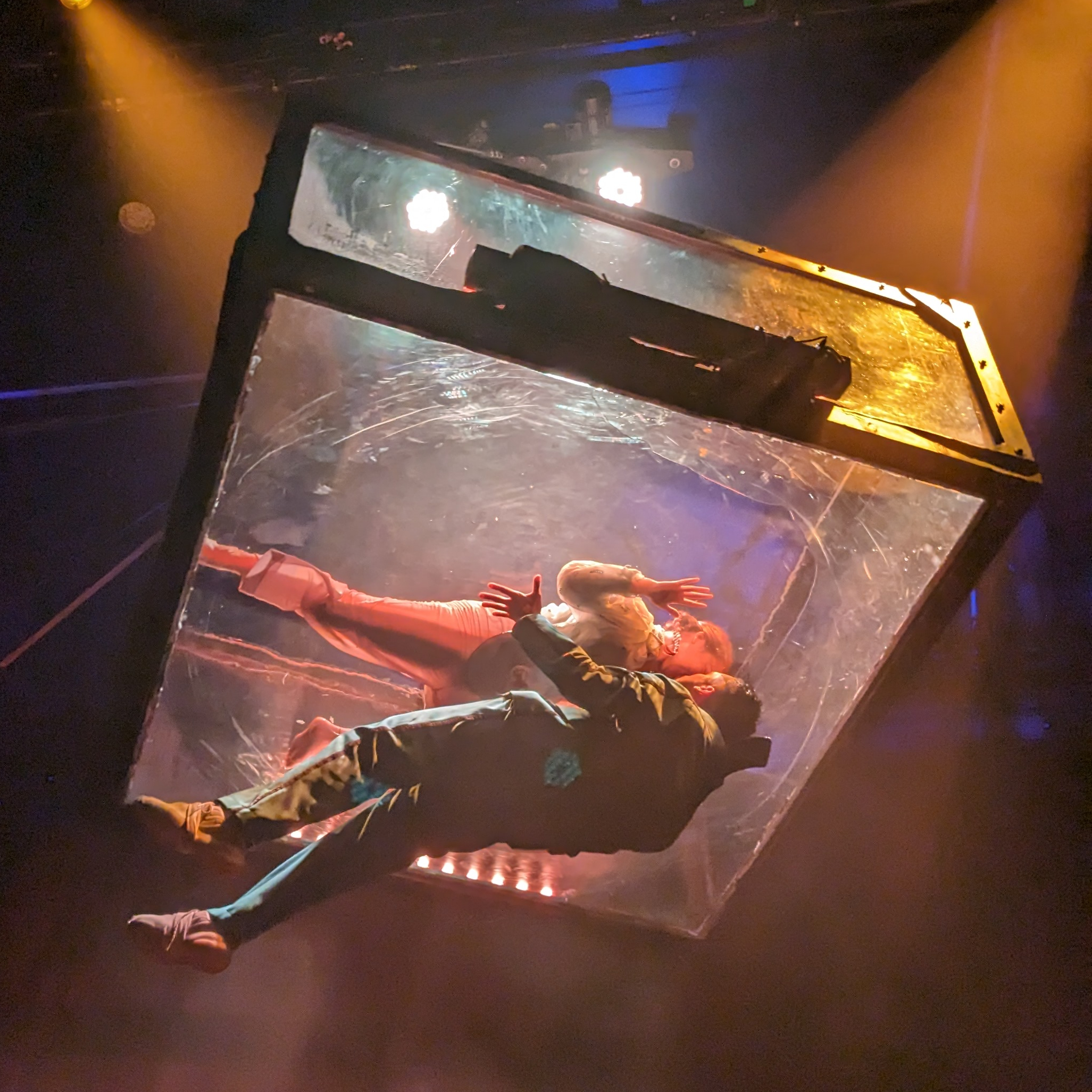 Clear box suspended above crowd with woman inside, pressing herself to the bottom of the box "kissing" a man suspended from the bottom of the box