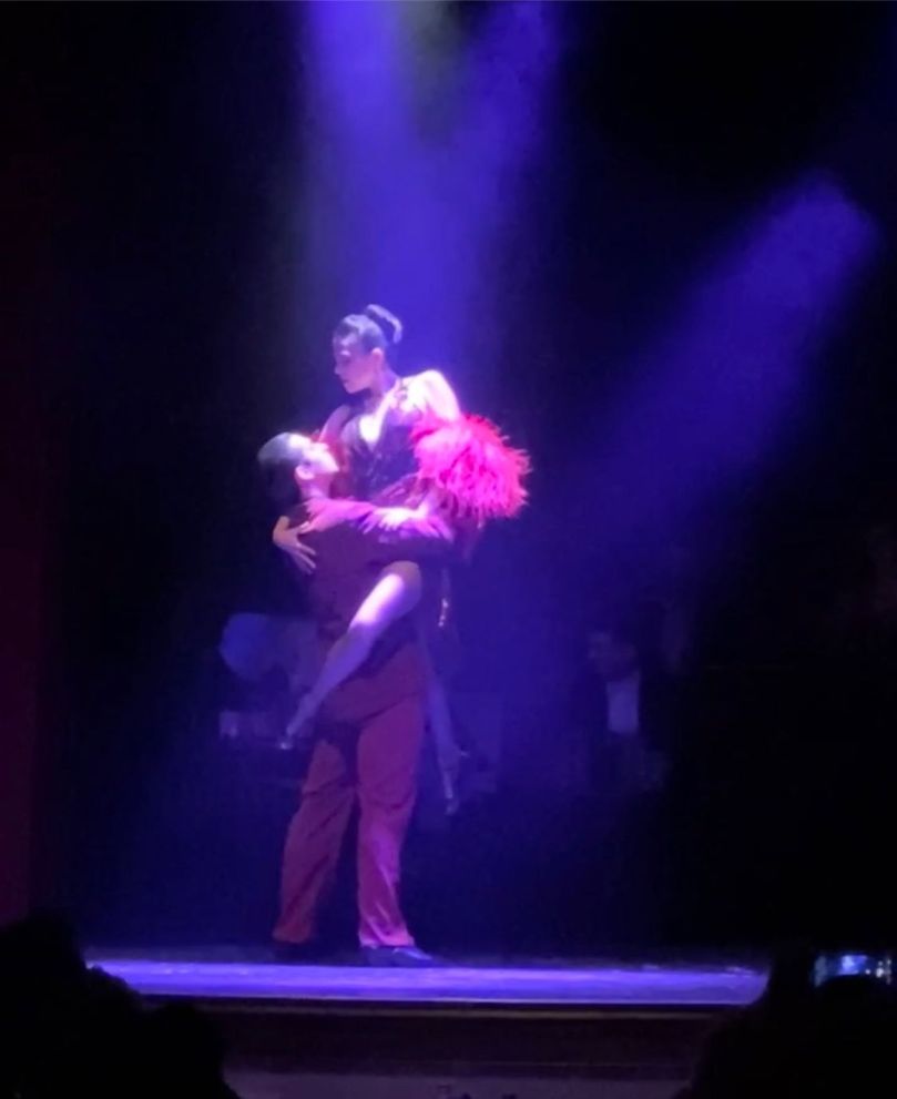 A couple dancing the Tango, dressed in red, with the man holding the woman off the ground, tightly, with her leg wrapped around his waist